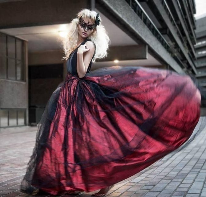 Red ballgown Blonde woman in a black eyemask wearing a red ballgown with black netting over the red. Street background.